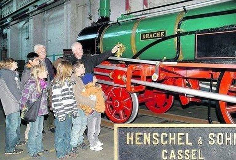 Technik ganz groß: Besonders beeindruckt zeigten sich die sechs bis zehnjährigen Hortkinder aus Vellmar-West vom Nachbau des Drachen, der ersten Henschel-Lokomotive, die 1848 gebaut wurde. Kurt Müller (links) und Museumsleiter Helmut Weich erklärten den Kindern alles Wissenswerte über Lok- und Lkw-Bau in der ehemaligen Kasseler Fabrik. Foto: Brandau